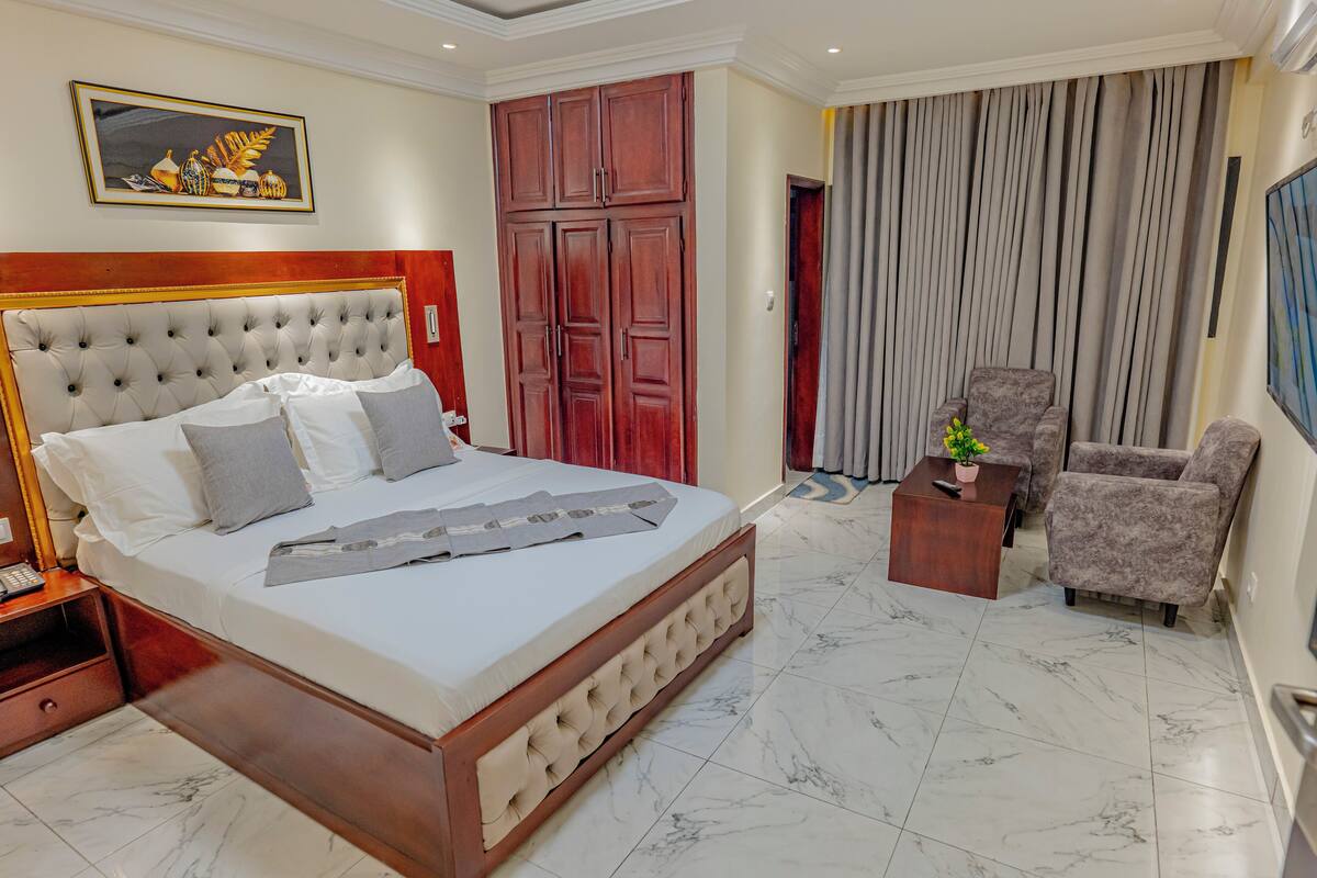 A hotel room with a large bed, a wooden headboard, a wardrobe, a seating area with a chair and a small table, and a painting on the wall.