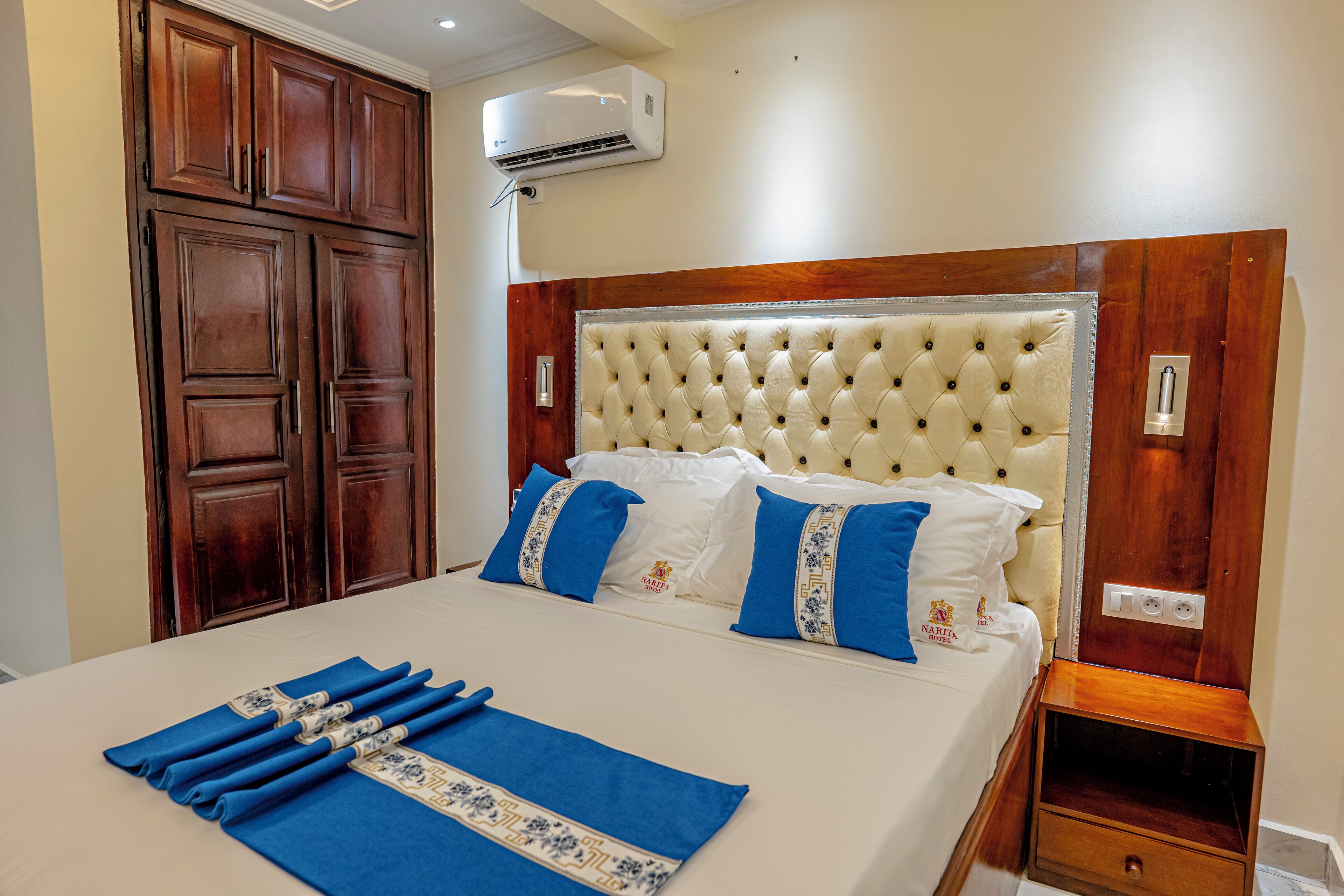 A neatly made bed with blue and white linens, a wooden wardrobe, and a wall-mounted air conditioner.