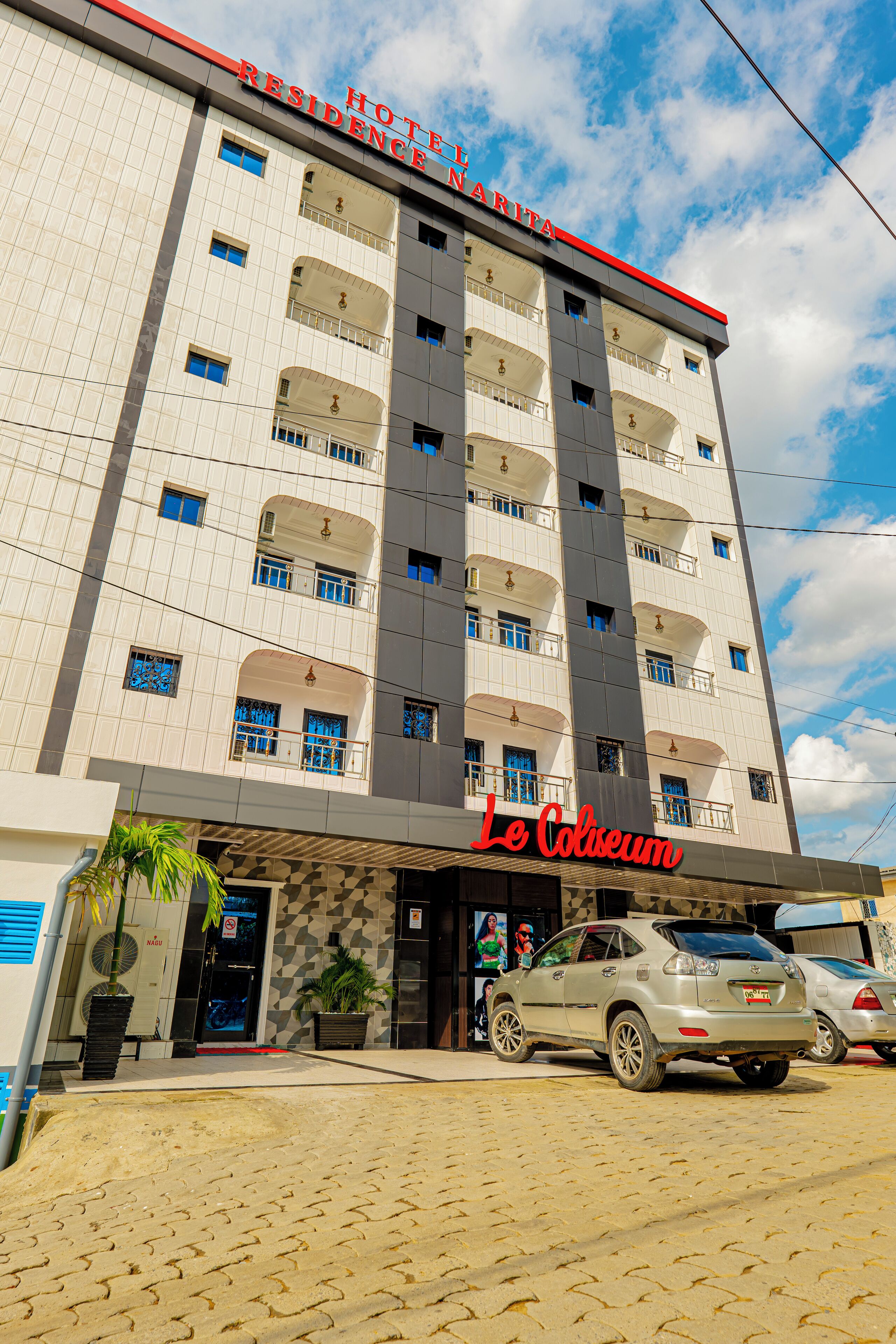 A hotel with multiple balconies, a prominent red sign, and a parking area with cars.