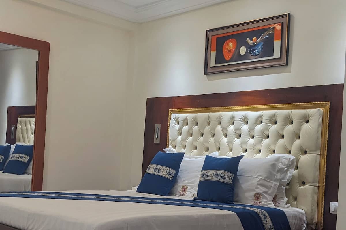 A bedroom with a large bed, a wooden headboard, a bedside table with a remote control, and a potted plant.