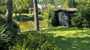 En rogivande utomhusmiljö med en damm, frodig grönska och en liten träbyggnad, troligen ett förråd eller en stuga, omgiven av träd och ett hus i bakgrunden.