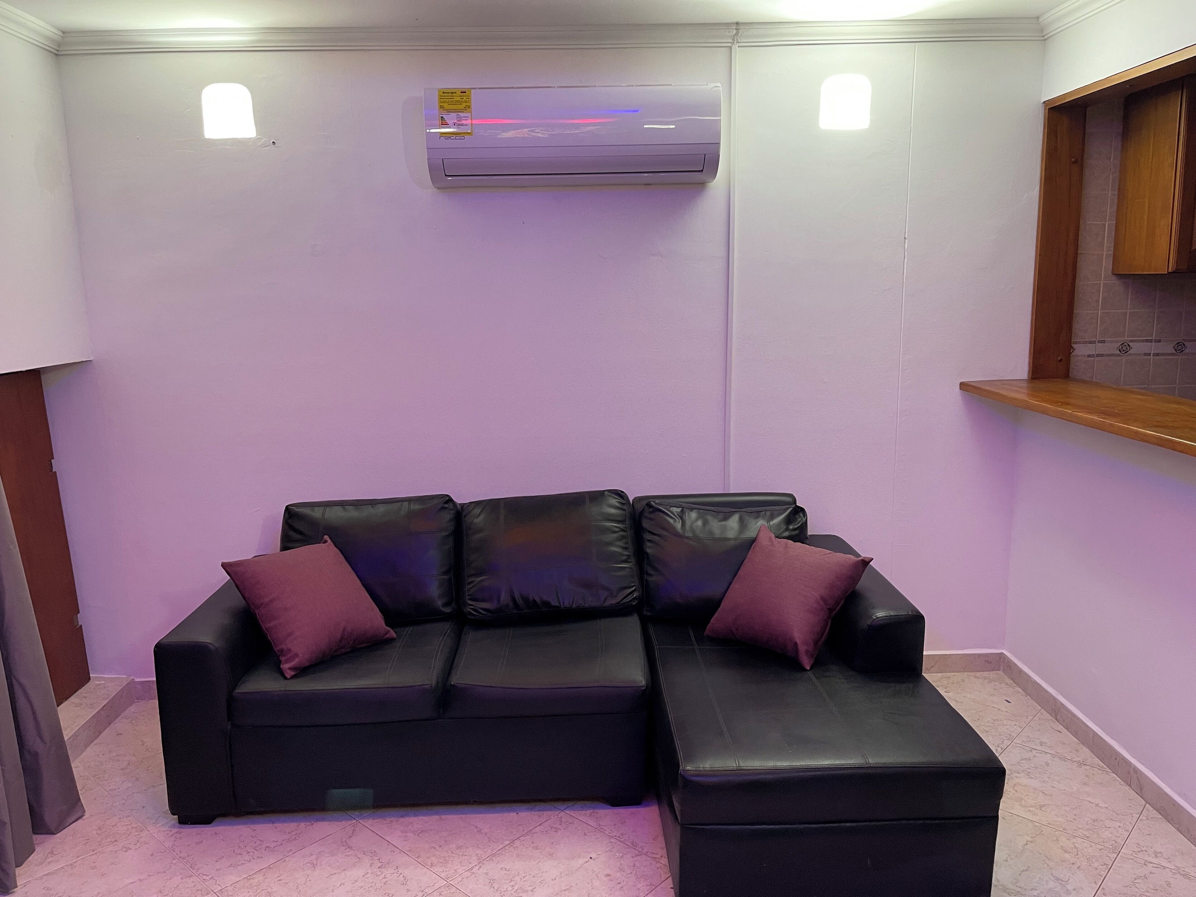 A room with a black leather sectional sofa with purple cushions, a small window, an air conditioning unit, a wooden shelf, and a tiled floor.