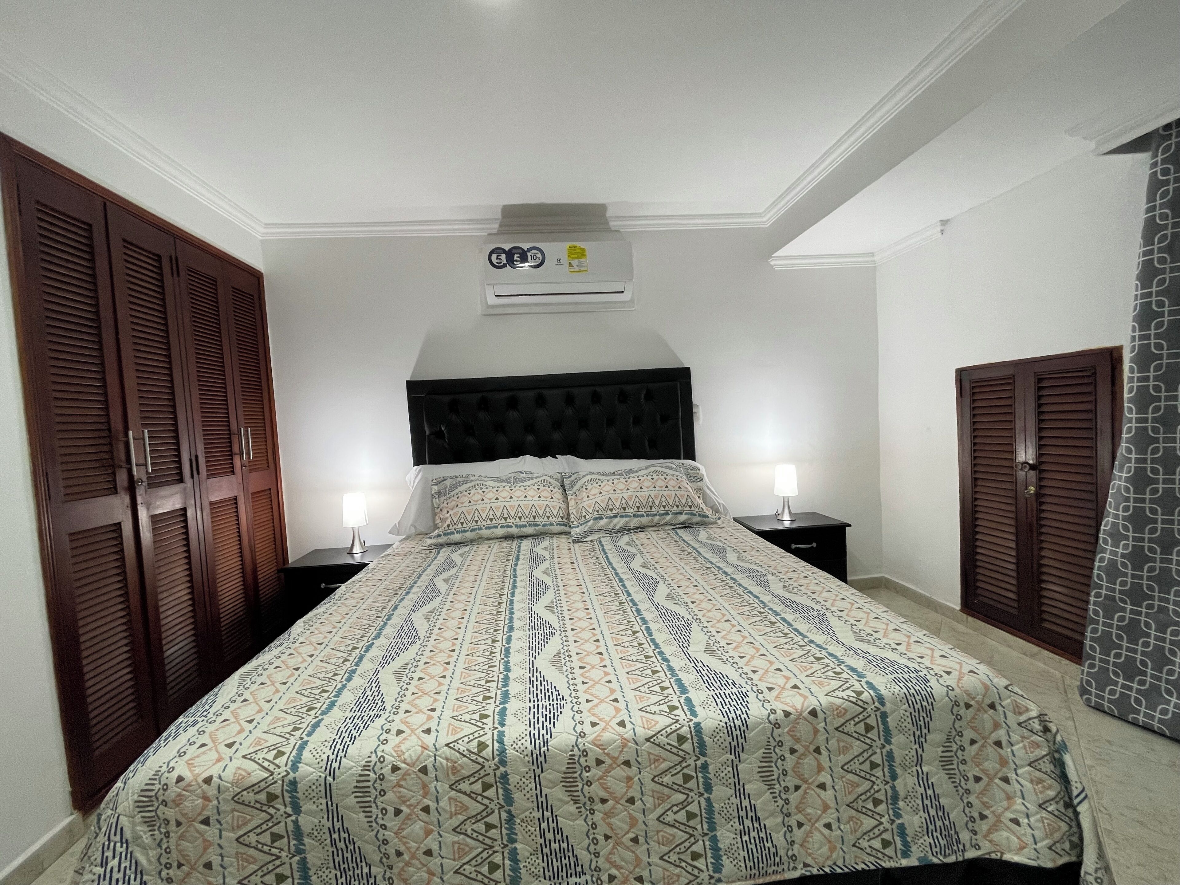 A bedroom with a bed, two bedside tables, a wall-mounted air conditioner, and a wardrobe.