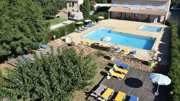Vue aérienne d’un complexe hôtelier doté d’une piscine entourée de chaises longues et de parasols, d’un petit bâtiment et d’une végétation luxuriante.