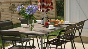 Un espace de restauration en plein air avec une table en bois, des chaises en métal, un parasol à rayures, un vase avec des fleurs violettes et des aliments pour le petit-déjeuner.