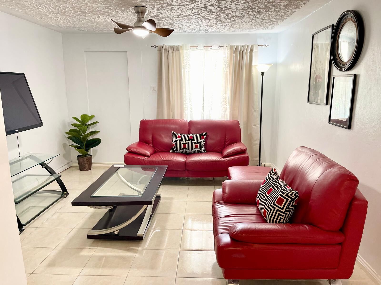 Uma sala de estar com sofá de couro vermelho, mesa de centro de vidro, ventilador de teto, planta em vaso e abajur.
