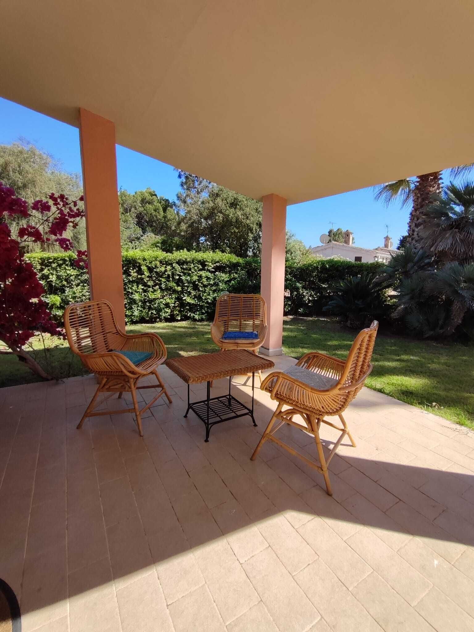 Un espacio de patio cubierto con mobiliario de mimbre, incluyendo sillas y una mesa, y vistas a un jardín exuberante y edificios al lejano.