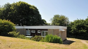 A small, single-story house with a brick exterior and white trim, surrounded by a grassy yard and dense trees. - 8 Hillside (Bude)