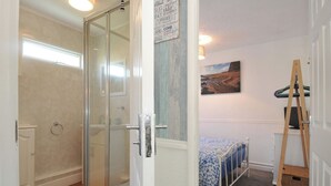 A room with a glass shower door, a bed with a blue and white patterned bedspread, a painting on the wall, and a wooden ladder leaning against the wall. - 8 Hillside (Bude)
