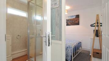 A room with a glass shower door, a bed with a blue and white patterned bedspread, a painting on the wall, and a wooden ladder leaning against the wall.
