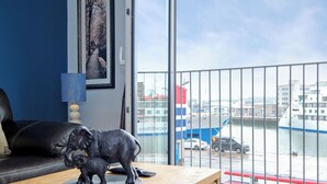 Un salon avec un canapé en cuir noir, une table basse en bois ornée d’une statuette, un tableau encadré au mur et une vue sur un port avec des bateaux et un navire.