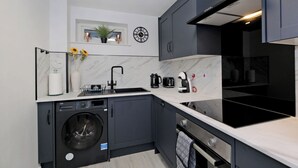 Une cuisine moderne équipée d’un lave-vaisselle, d’un lave-linge, de meubles de couleur noire, d’un plan de travail blanc, d’une horloge et d’une petite plante.