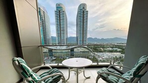 Ein Balkon mit zwei gestreiften Liegestühlen, einem kleinen runden Tisch und Blick auf zwei hohe Gebäude und entfernte Berge.