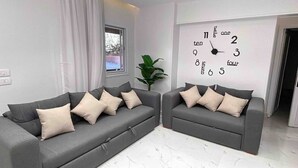 A modern living room with two grey sofas, a large wall clock, and a window with curtains. - Your Unique home in cairo (Cairo)