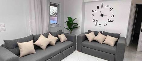 Un salon moderne avec deux canapés gris, une grande horloge murale et une fenêtre avec des rideaux.