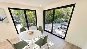 A modern dining area with a round table and six chairs, large windows offering a view of an outdoor patio with a table and chairs. - Large 4Br/3Bh Beach House, Bikes,  Chairs & More (Sanibel)
