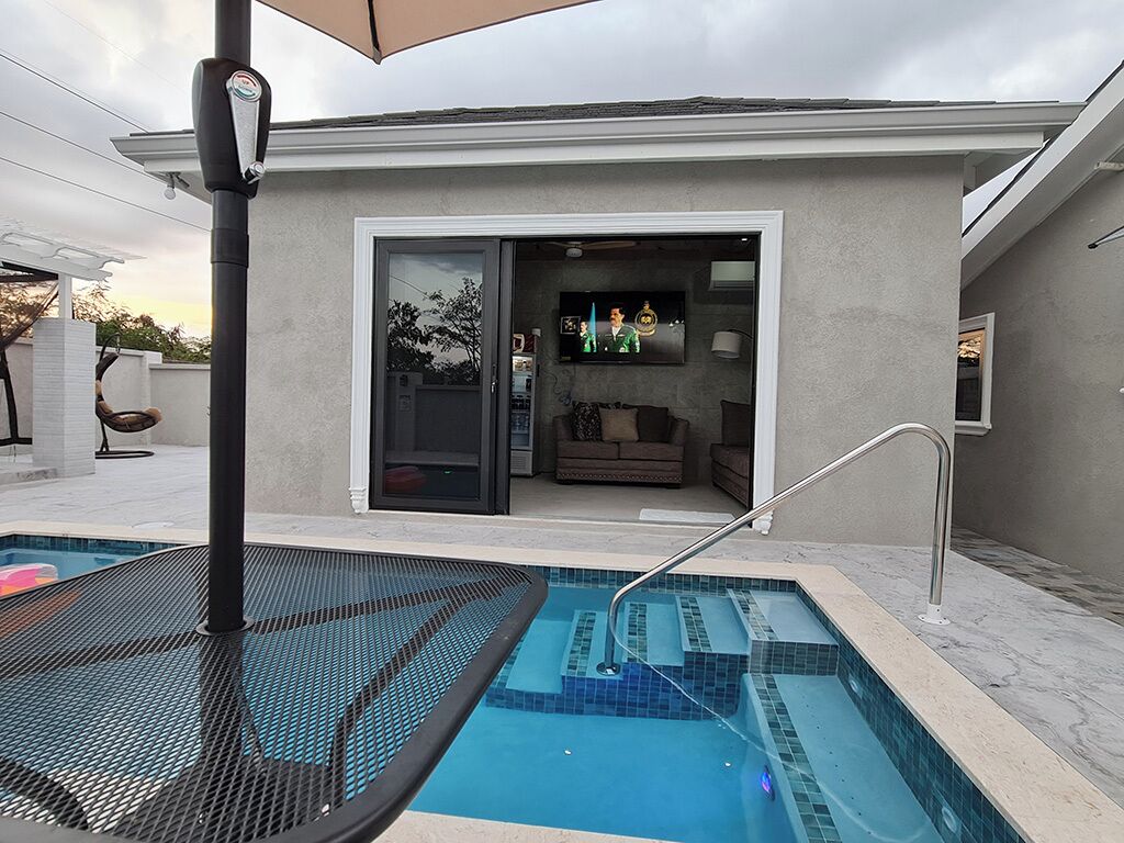 Ein moderner Poolbereich im Freien mit Glastür zu einem Wohnzimmer, einem Metalltisch und einer Liege am Pool.