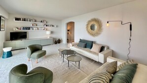 A modern living room with a sofa, armchairs, a coffee table, and a TV. There are bookshelves and a decorative mirror on the wall. - Apartment facing the sea (Ajaccio)