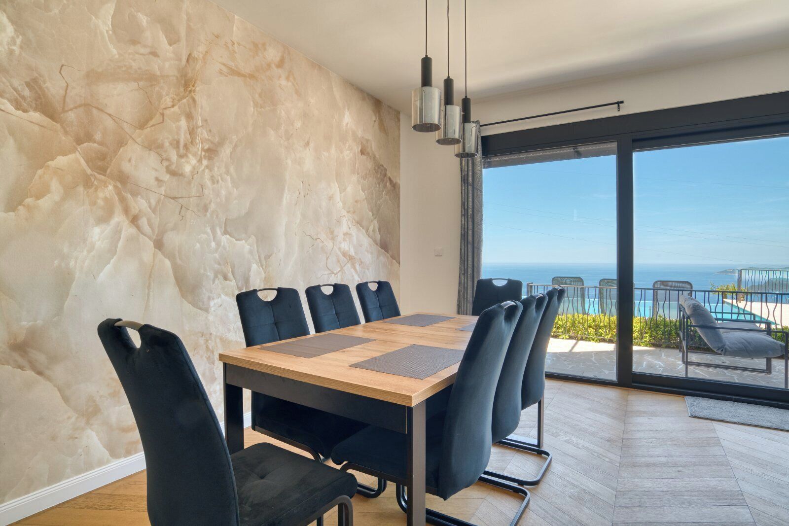 Un espace repas agrémenté d’un mur en marbre, d’une table en bois, de chaises noires et offrant une vue sur l’océan grâce à de grandes portes vitrées.