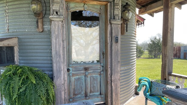 A rustic wooden building with a weathered door, a porch with a bench, and a metal railing. - 8 Minutes from Round Top!  Your Silo-Bration Destination
StoneHENge Silo #2 (La Grange)