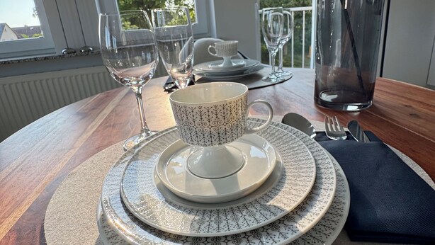 A dining table set with plates, a cup, and cutlery, with a view of trees and a balcony in the background.