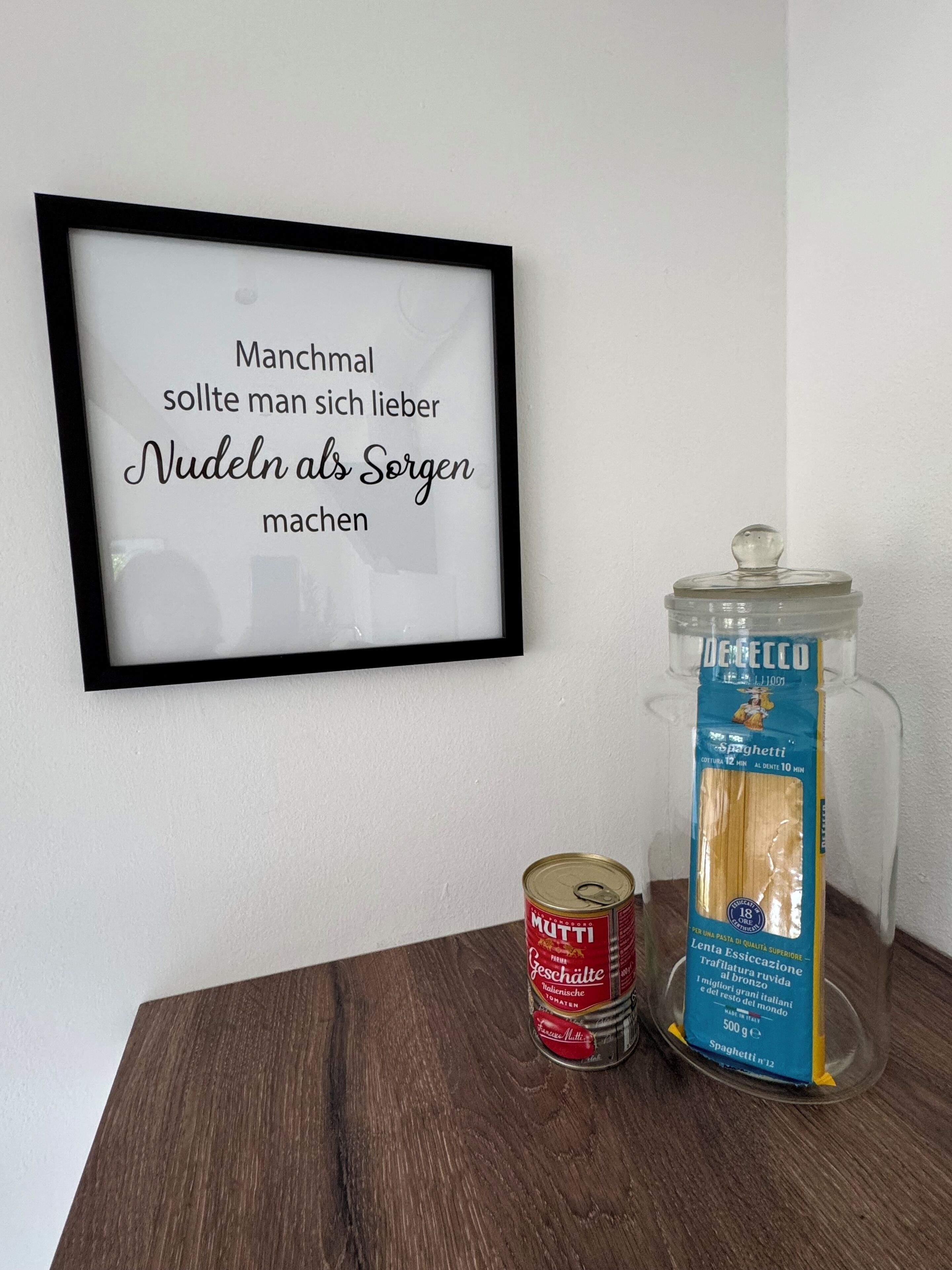 A framed quote on a wall, a can of Muttis, and a package of De Cecco spaghetti.