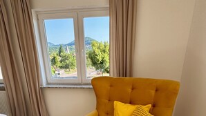A yellow sofa bed with matching cushions in a room with a large window, beige curtains, and a view of greenery and hills.