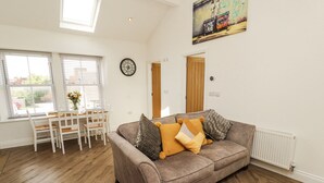 A living room with a sofa, a dining table with chairs, a clock, and a radiator. - Lakes View Apartment 2 (Carnforth)