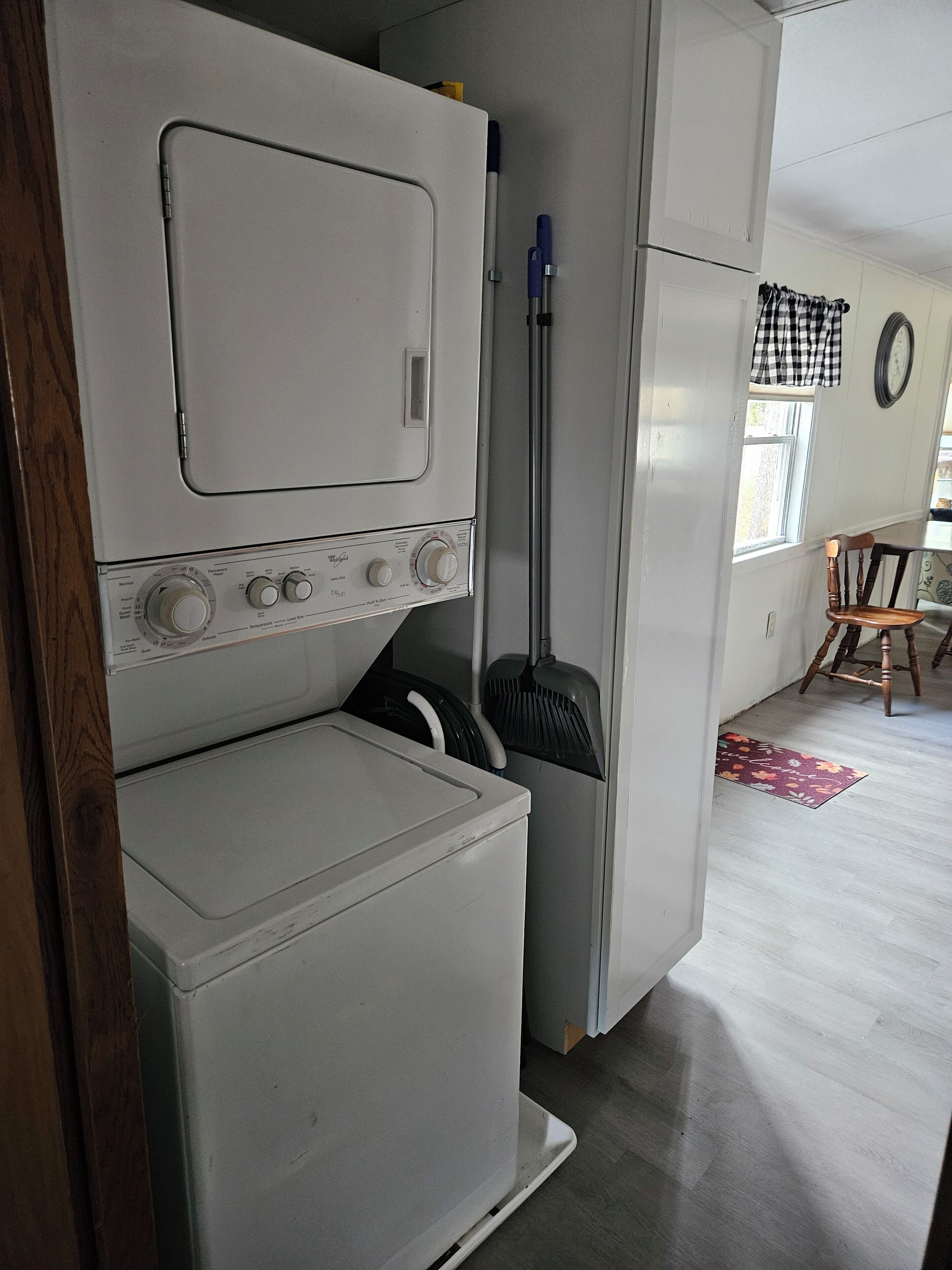 A compact laundry area with a washing machine and dryer, a broom, and a window with a checkered curtain.