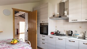 Een moderne keuken met witte kastjes, een gasfornuis en een magnetron. Er is een eettafel met een tafelkleed met bloemenprint en een fruitschaal.