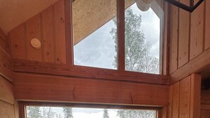 Un interior de madera con un ventanal grande, un sofá con cojines estampados y vistas a un paisaje verde.