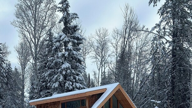 Una cabaña de madera con techo inclinado, ventanas amplias y un paisaje cubierto de nieve.