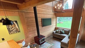 Un acogedor interior de cabaña con una mesa de comedor de madera, sillas, un sofá, una chimenea y un ventanal que ofrece vistas al exterior.