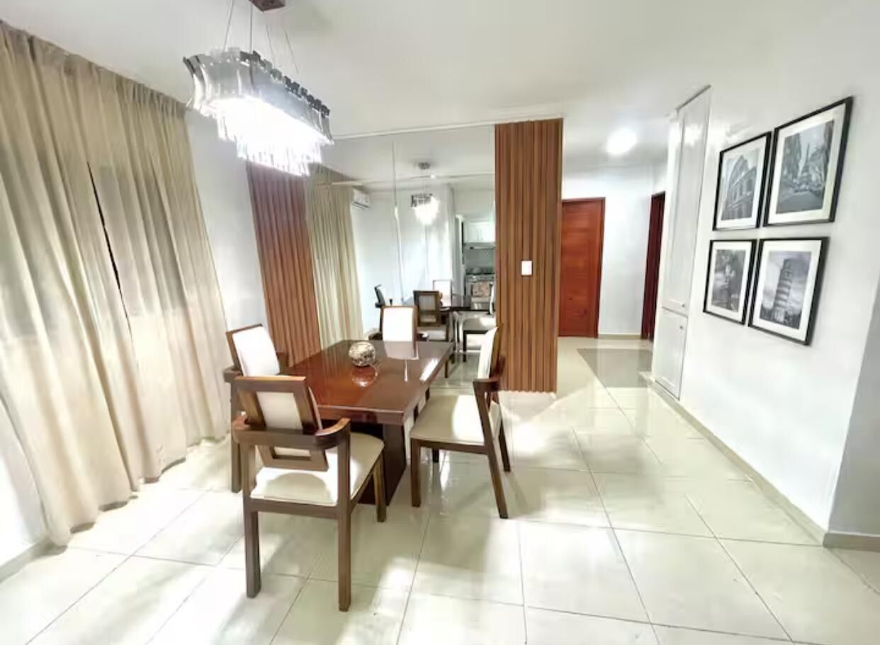 Un comedor con una mesa y sillas de madera, una lámpara de araña y una cocina al fondo.