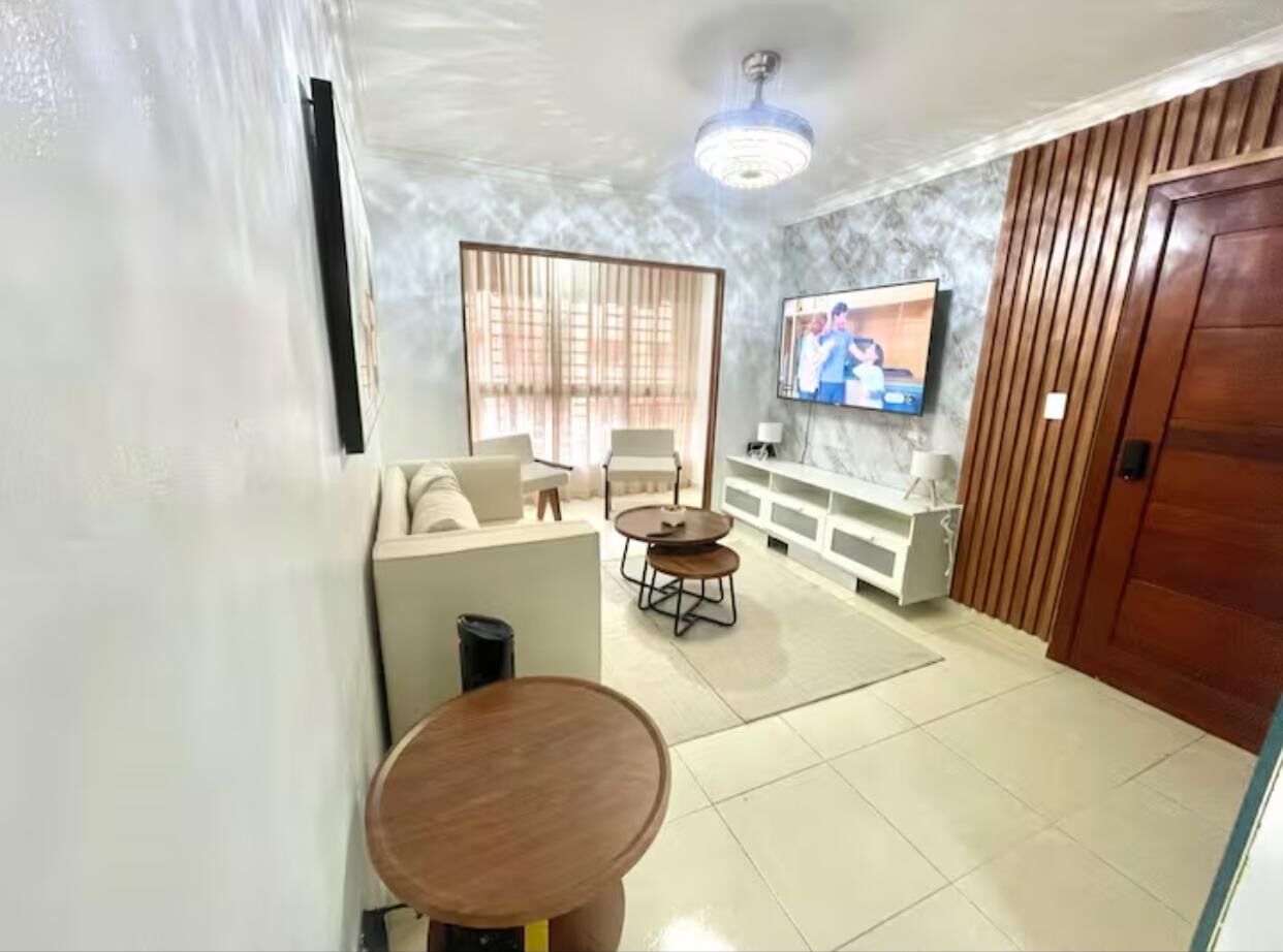 A living room with a sofa set, two round tables, a TV mounted on the wall, and a wooden door.
