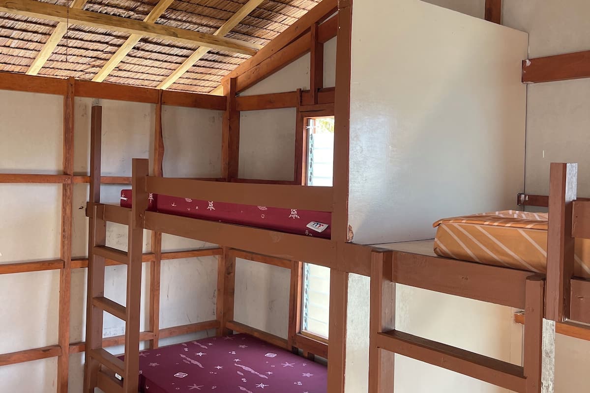 Une chambre avec des lits superposés, un ventilateur de plafond, des poutres en bois et un toit de chaume.