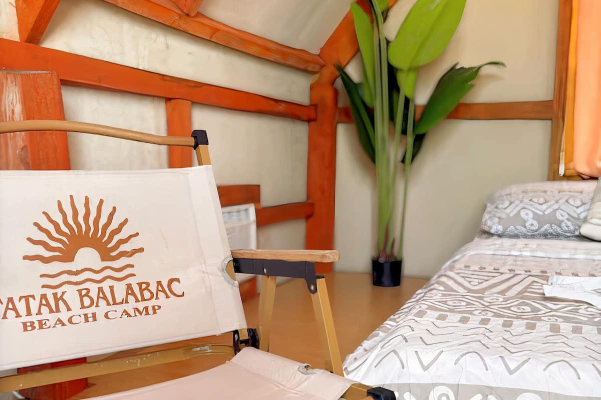 Une chambre d’un campement de plage, comprenant un lit, une chaise, une grande plante et un mur orné d’un logo représentant un soleil.