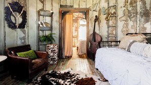 Una habitación rústica con estética vintage, que incluye un tapete de cuero, un sillón de cuero, una cama con edredón blanco, una estantería de madera con objetos decorativos y un umbral que da a otra habitación.