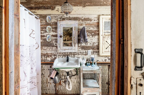 Un baño rústico con un lavamanos antiguo, ducha suspendida y pared de madera con pintura descascarada.