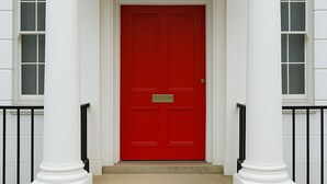 Una entrada de edificio con una puerta roja, columnas blancas y una barandilla negra.
