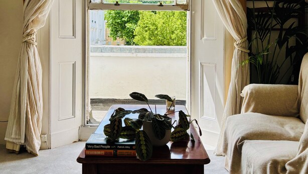 Una habitación con un ventanal grande, una mesa con libros, una silla y una planta en maceta.