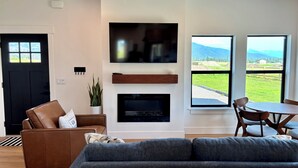 Una sala moderna con chimenea, televisor, sofá, mesa de comedor con sillas y ventanas amplias con vistas al exterior.