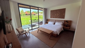 A modern hotel room with a large bed, a desk, and a chair. The room has a large window offering a view of a lush garden and a building. - Villa Rizière Ubud Center (Bali)