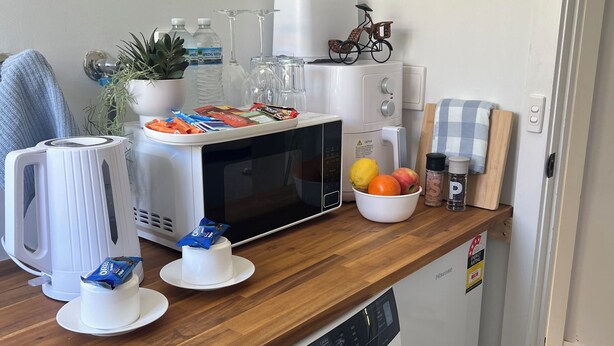 Un espacio tipo cocina con microondas, hervidor y lavadora. Hay dos pasteles pequeños en la encimera, un cuenco de fruta y un sombrero en la pared.