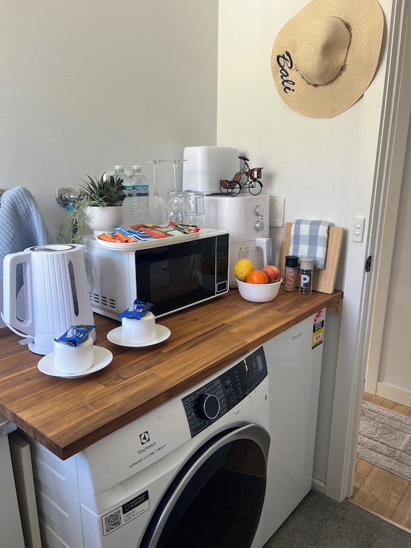 Un espacio tipo cocina con microondas, hervidor y lavadora. Hay dos pasteles pequeños en la encimera, un cuenco de fruta y un sombrero en la pared.