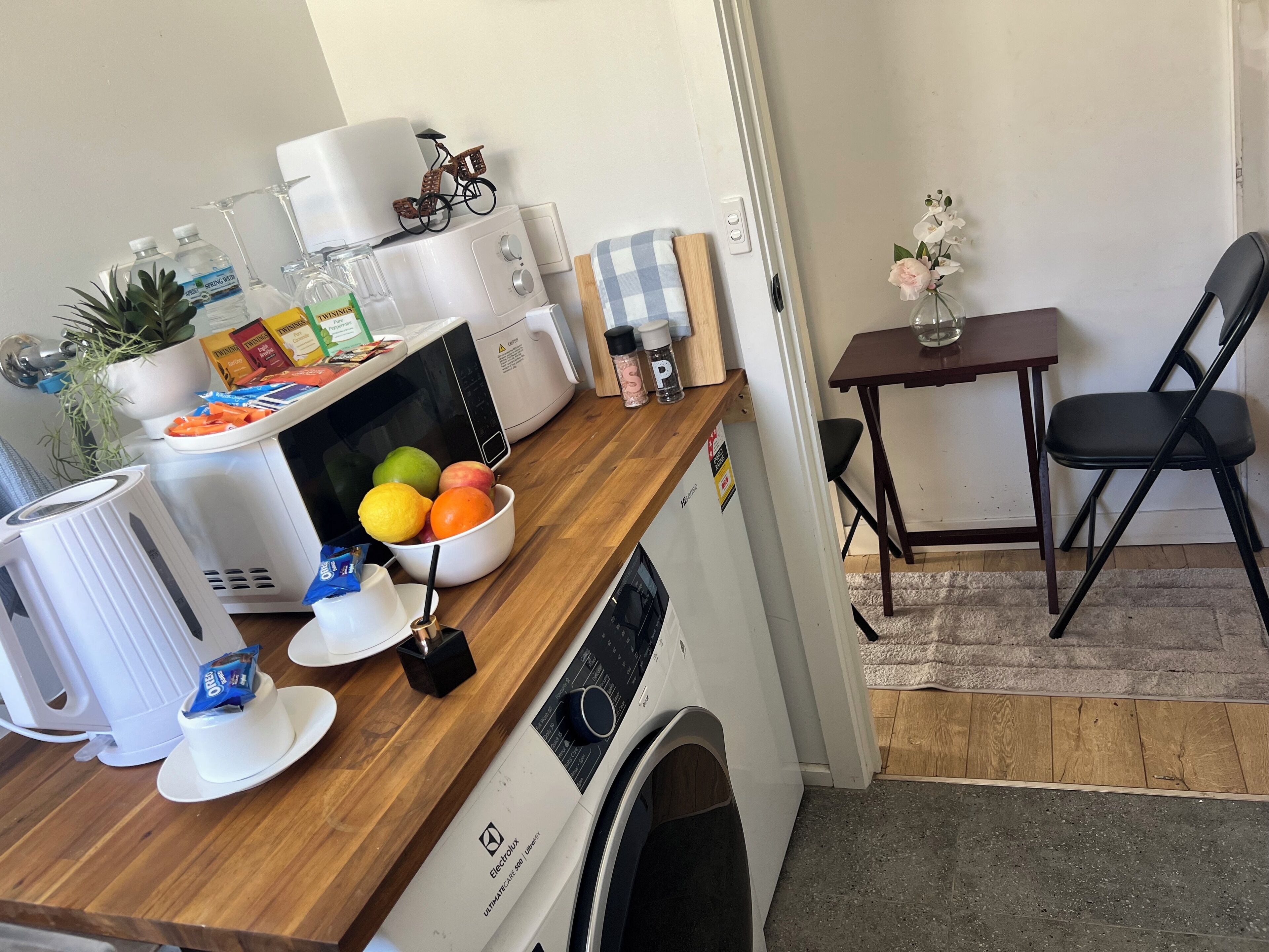 A compact kitchen with a microwave, toaster, and coffee maker. There is a bowl of fruit on the counter and a small table with a chair.