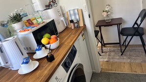 Cocina compacta con microondas, tostadora y cafetera. Hay un cuenco de fruta en la encimera y una pequeña mesa con una silla.