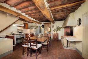 A rustic kitchen with exposed wooden beams, a stone fireplace, and a dining area with wooden chairs and a table.
