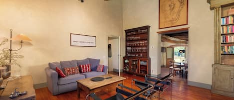 Een ruime woonkamer met een houten plafond, een bank, een bijzettafeltje, een boekenkast en een eethoek met stoelen.