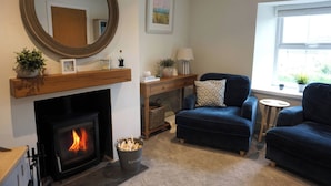 A cozy living room with a fireplace, a round mirror, a blue armchair, a small table, and a window with a view. - Glendale (Port Isaac)